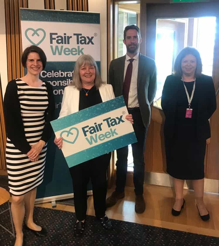 four people standing in front of Fair Tax Week banner, and woman in the middle holding Fair Tax Week photo prop
